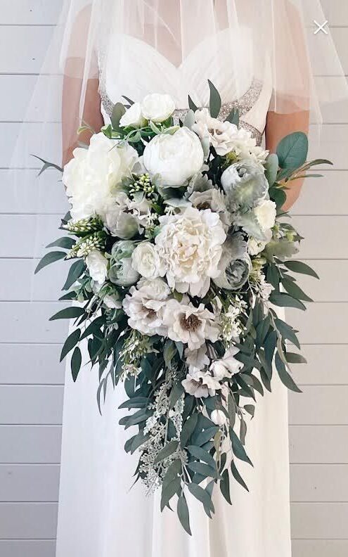 A cascading bridal bouquet featuring large white peonies and trailing eucalyptus greenery against a white wooden wall.