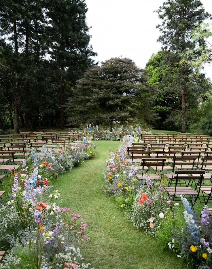 Rows of wooden ceremony chairs set outdoors on a lawn, with vibrant clusters of wildflowers appearing to grow naturally from the grass at the end of each row.