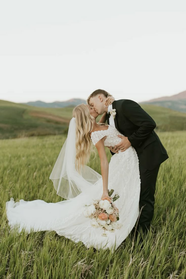 Groom dipping bride for a kiss in a green field with mountains, bride wearing long veil and holding bouquet.