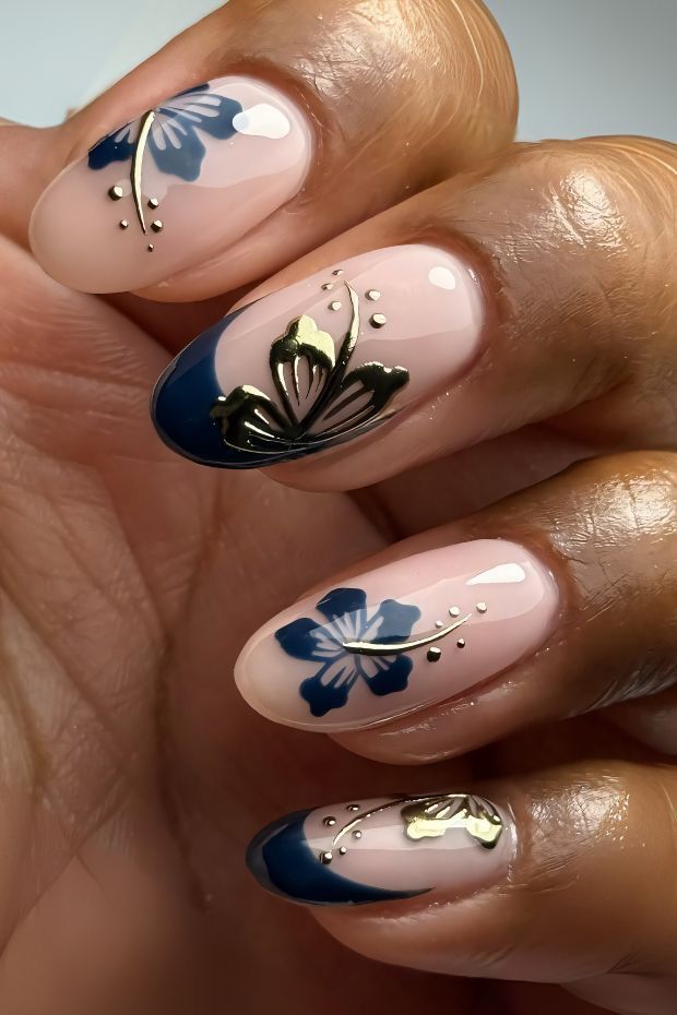 Close up of oval shaped nails with a glossy nude base, decorated with deep blue hibiscus flowers and metallic gold stems and dots.