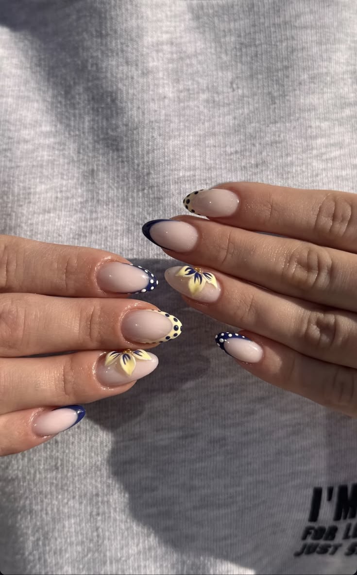 Almond shaped nails with a nude base and deep navy blue French tips, featuring white polka dots on the tips and 3D yellow hibiscus flower accents.
