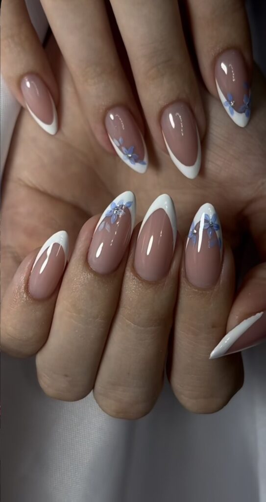 Long pointed almond shaped nails with a clean nude base and classic white French tips, decorated with delicate light blue flowers and tiny sparkling rhinestones.