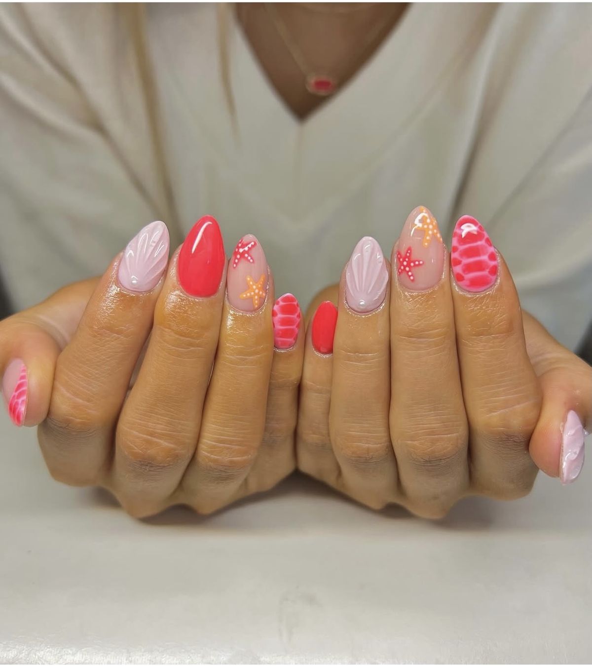 Almond shaped nails with a mix of coral red polish, pink 3D shell textures, and simple white sea life drawings.