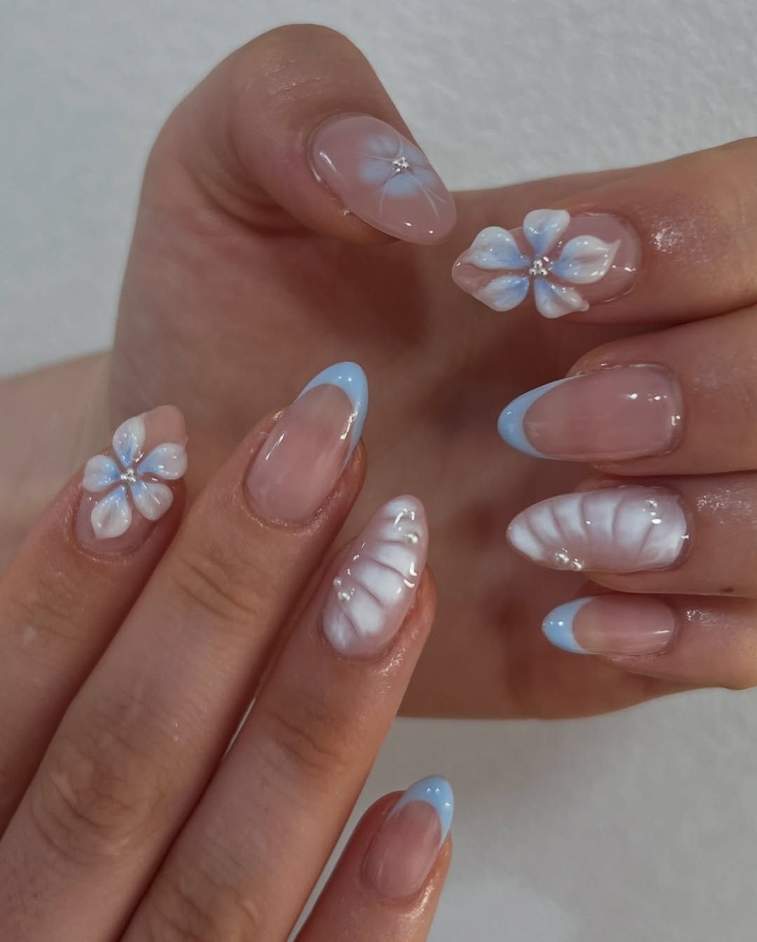 Long almond shaped nails with baby blue French tips, glossy pearl details, and clear accent nails with raised 3D white flowers.