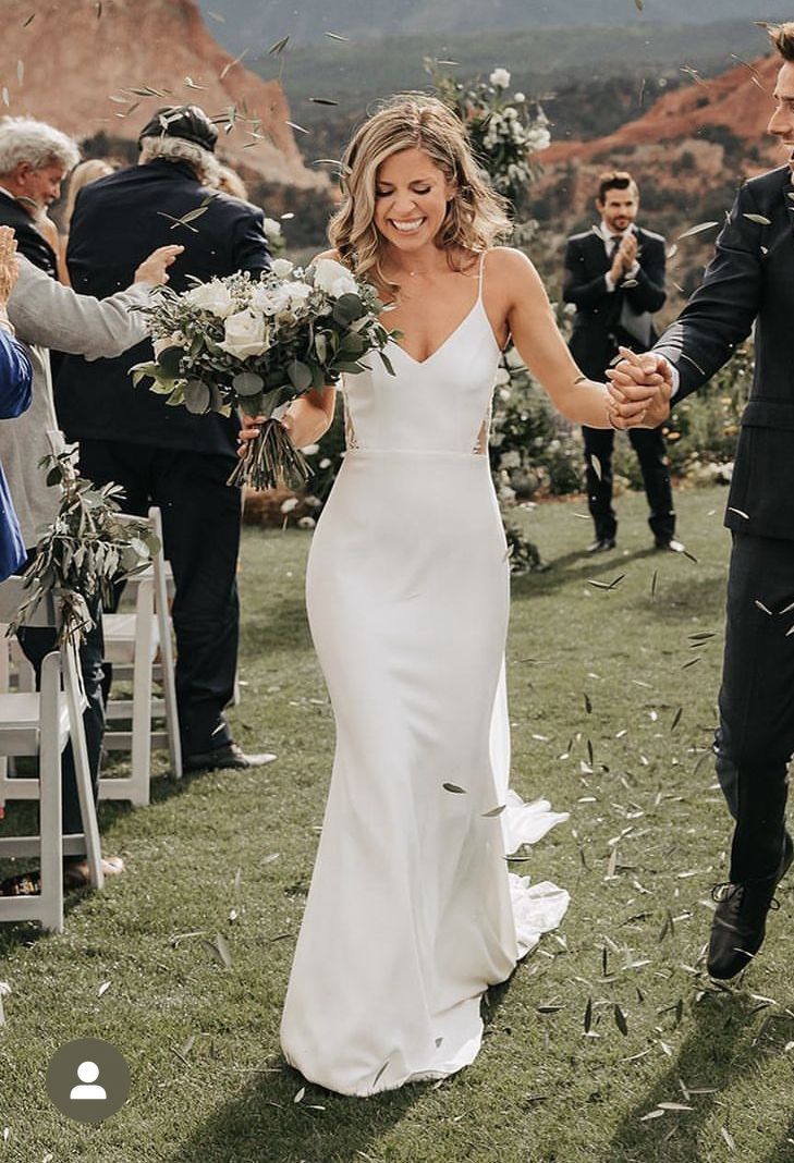 Bride wearing a minimalist white crepe column wedding dress with spaghetti straps and side cutouts walking through an outdoor ceremony.
