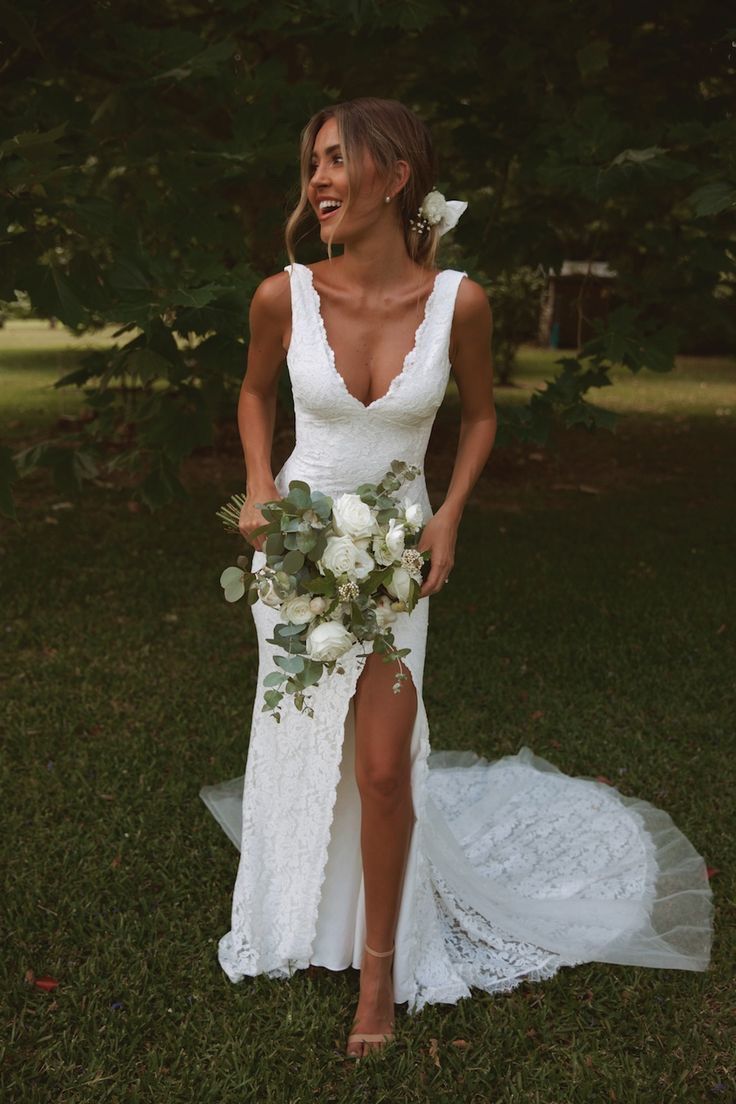 Bride smiling in a garden wearing a white lace wedding dress with a high leg slit and a deep V neckline.
