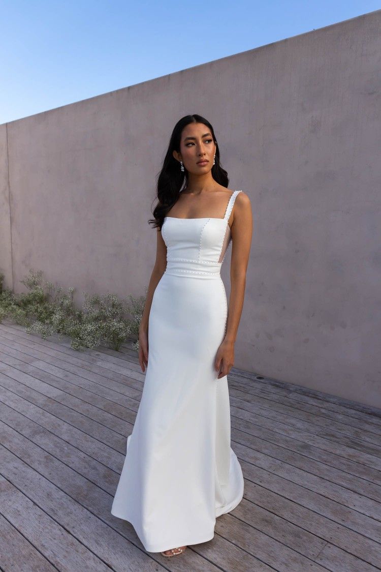 Bride standing outdoors against a concrete wall wearing a structured white square-neck wedding gown with textured straps.