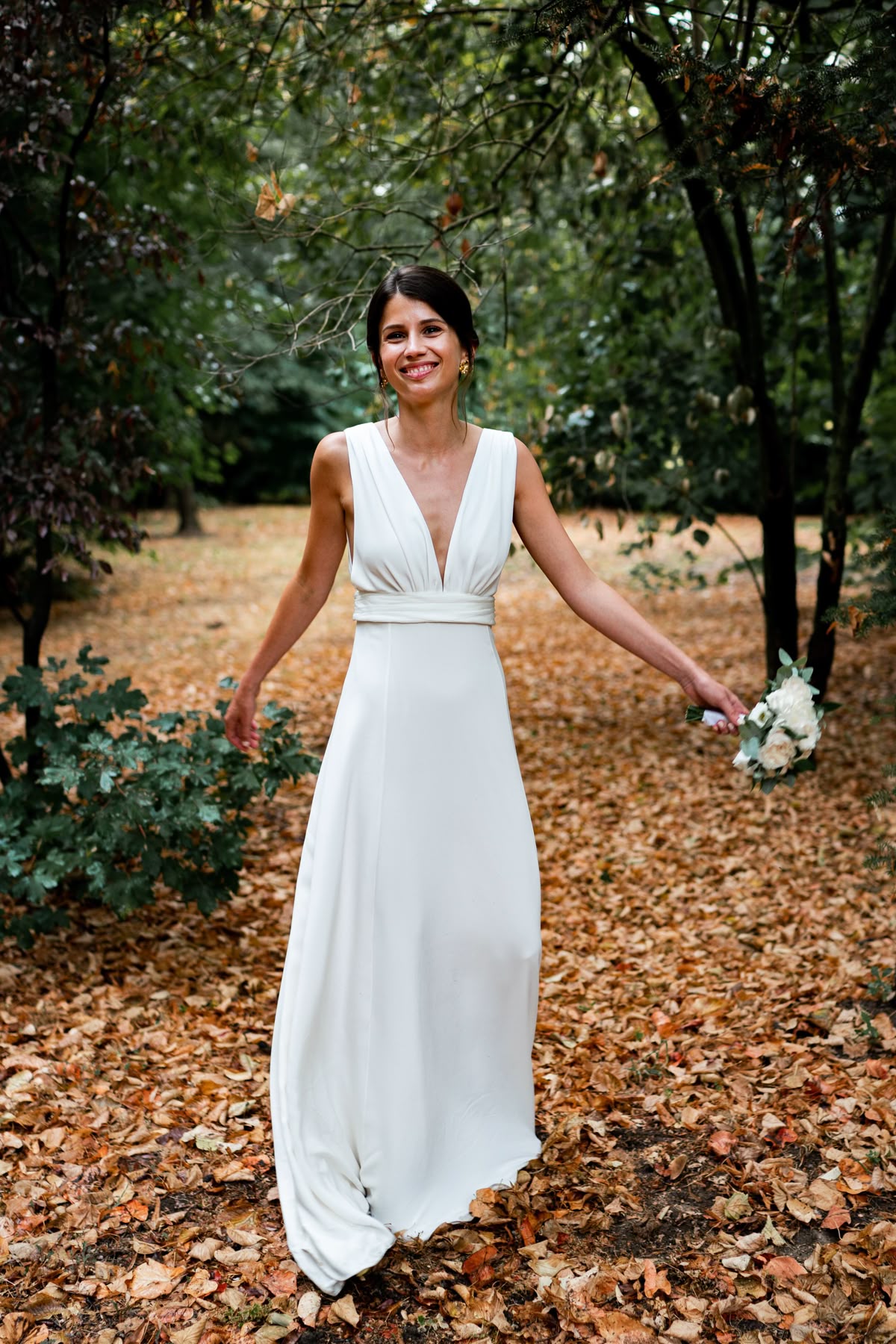 Bride smiling in a park filled with autumn leaves, wearing a flowing white empire-line wedding dress with a plunged bodice.