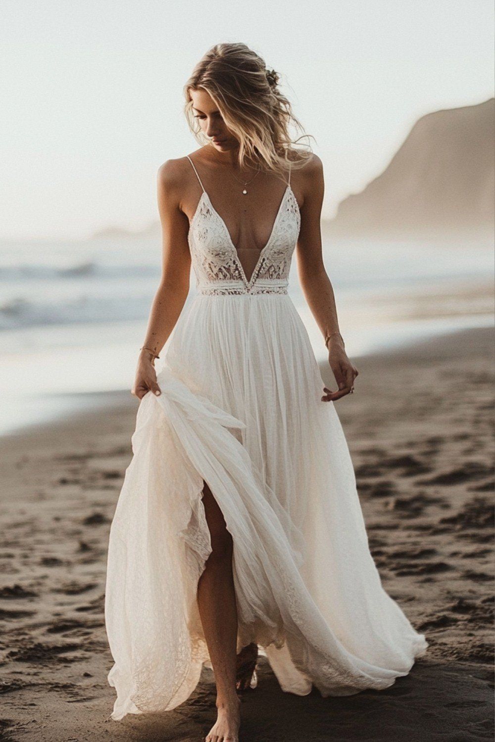 Bride walking on a beach wearing a wedding dress with a sheer, plunged lace bodice and a flowing white chiffon skirt with a slit.