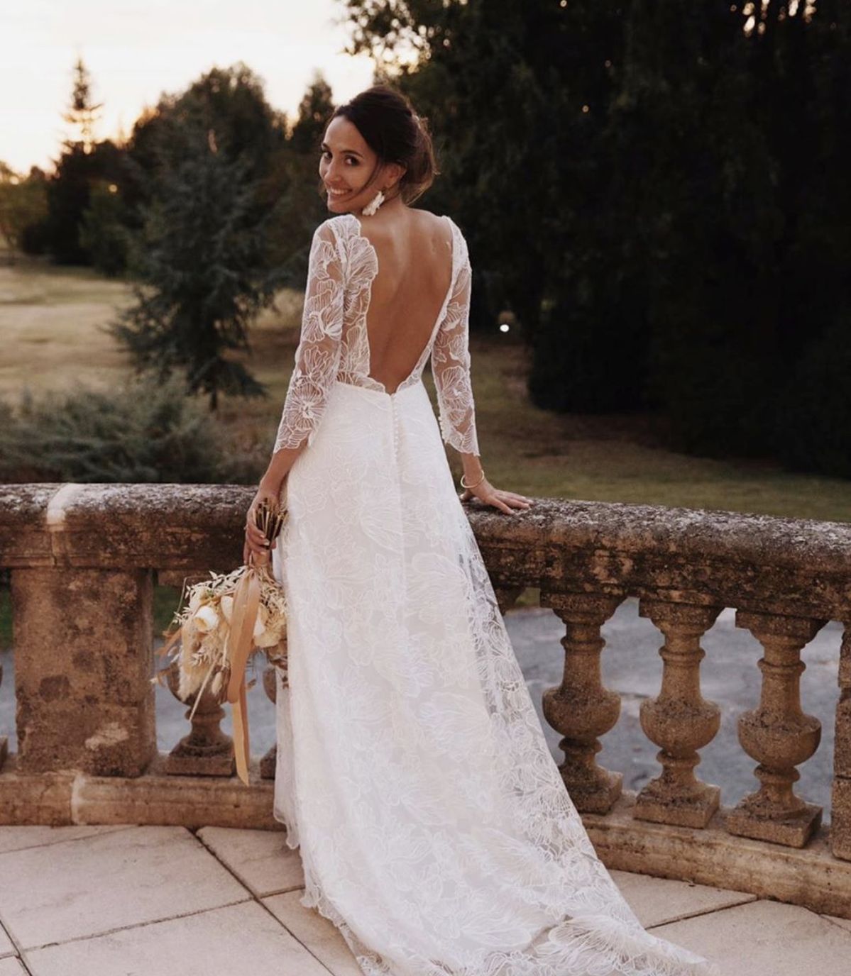 Back view of a bride looking back on a stone terrace, wearing a white long-sleeve wedding dress with an open back and illusion lace.
