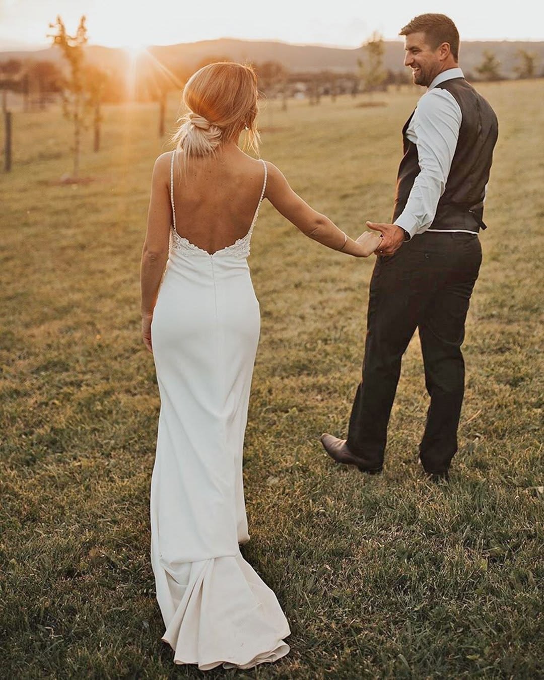 Back view of a bride walking with her groom at sunset, wearing a low-back wedding dress with beaded straps.