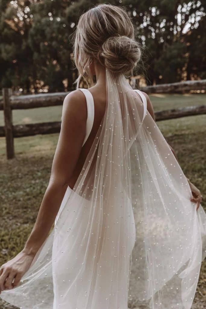 Back view of a bride with a low bun hairstyle wearing a pearl-embellished wedding veil over a simple low-back dress.