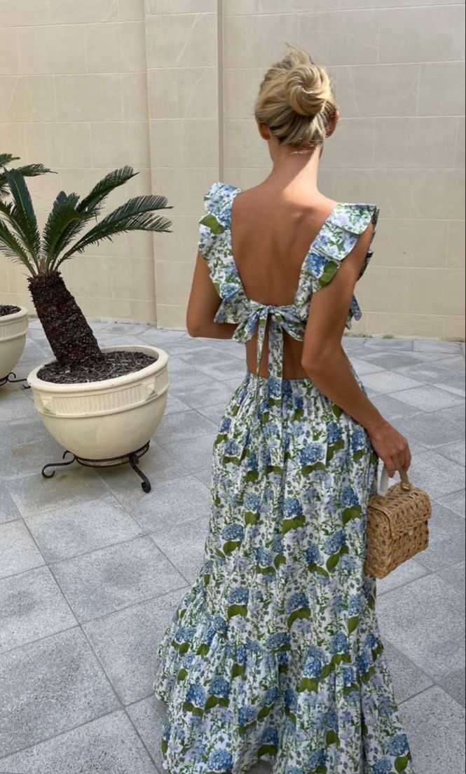 The back view of a woman with blonde hair in a bun wearing a blue hydrangea print maxi dress with an open tie-back design.