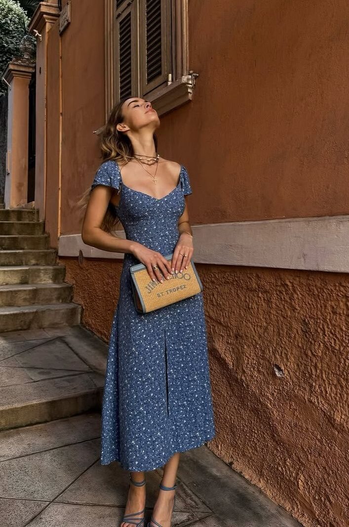 A woman standing beside an orange wall wearing a blue floral midi dress with cap sleeves and holding a woven straw clutch.