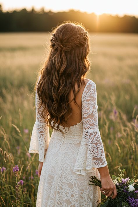 Back view of a bride with long wavy hair in a lace dress with bell sleeves in a field.