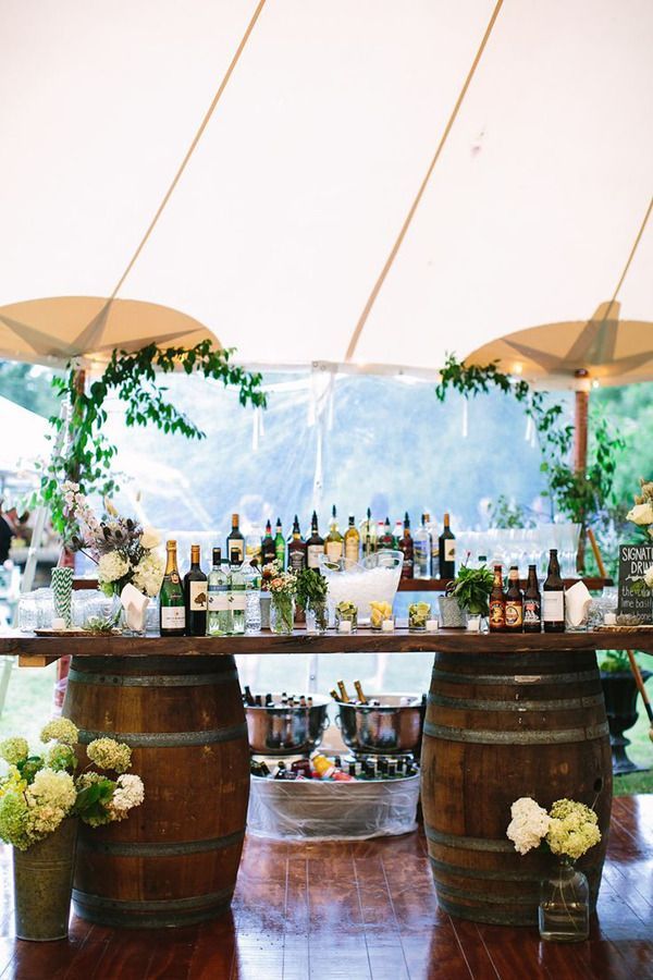 A rustic wedding bar under a tent made of a thick wood slab sitting on two wine barrels filled with liquor bottles and champagne.