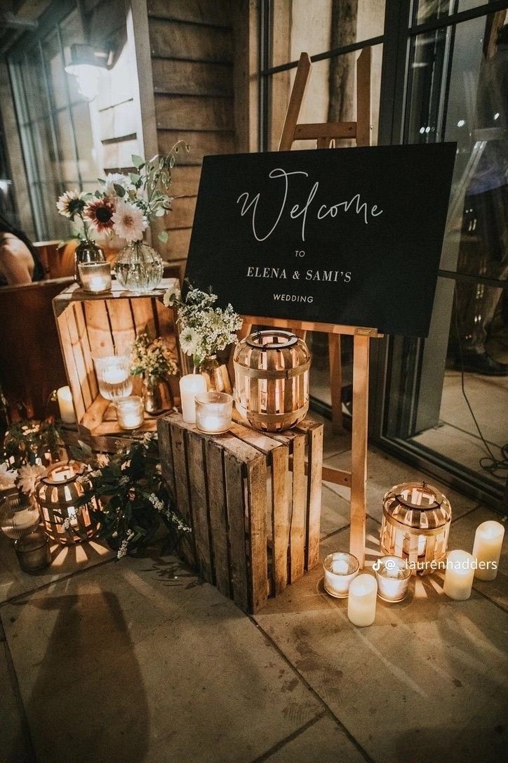 A cozy indoor wedding welcome sign on a wooden easel surrounded by stacked crates, glowing white candles, and wooden lanterns.