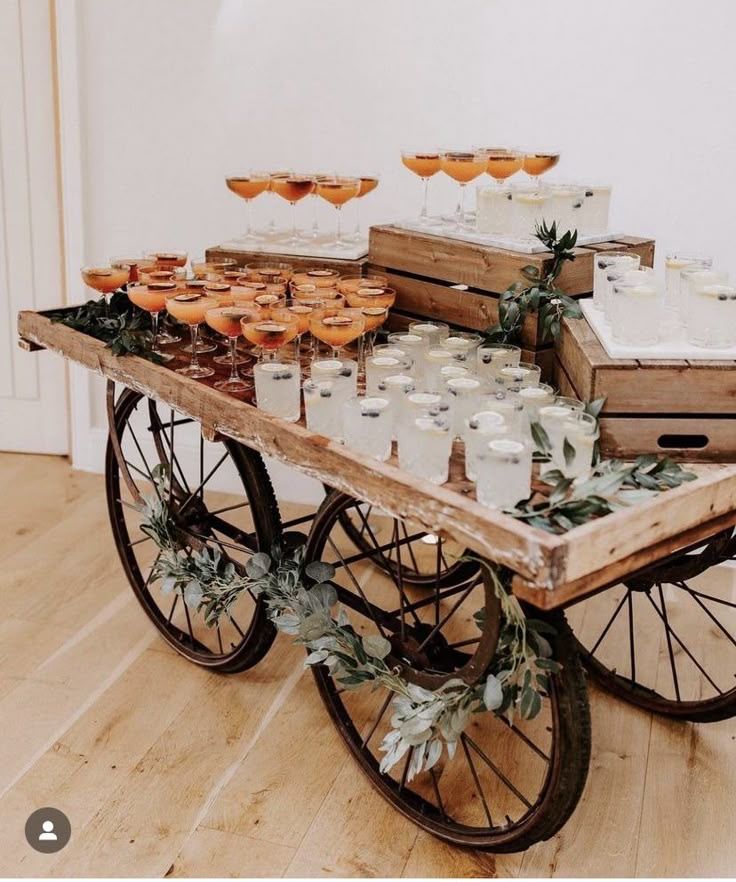 A vintage wooden cart on metal wheels holding rows of orange and clear signature cocktails on top of stacked wooden crates.
