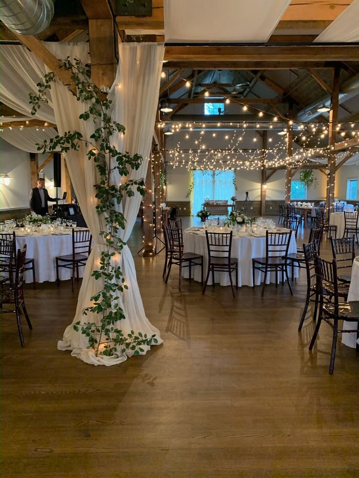 A large indoor wooden barn pillar wrapped in sheer white fabric, green leaves, and fairy lights near reception tables.