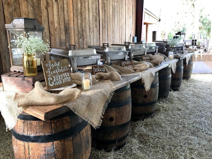 A long wedding buffet table supported by four wooden wine barrels with silver chafing dishes and a hessian runner inside a barn.