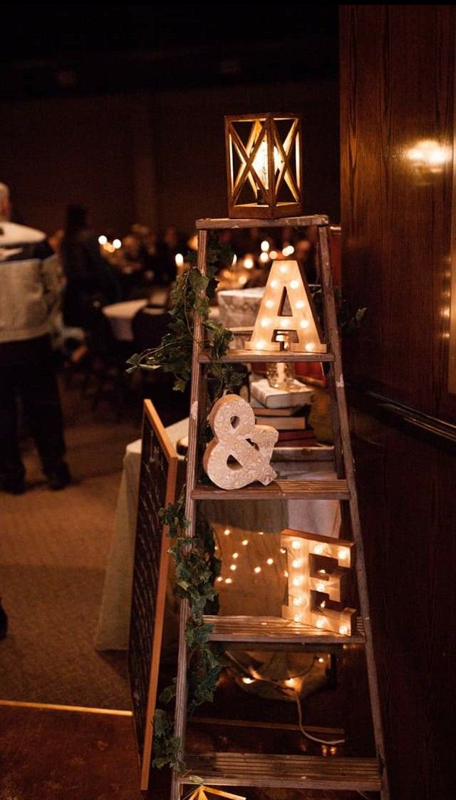 A vintage wooden stepladder inside a wedding reception holding bright glowing marquee letters and wrapped in green ivy leaves.
