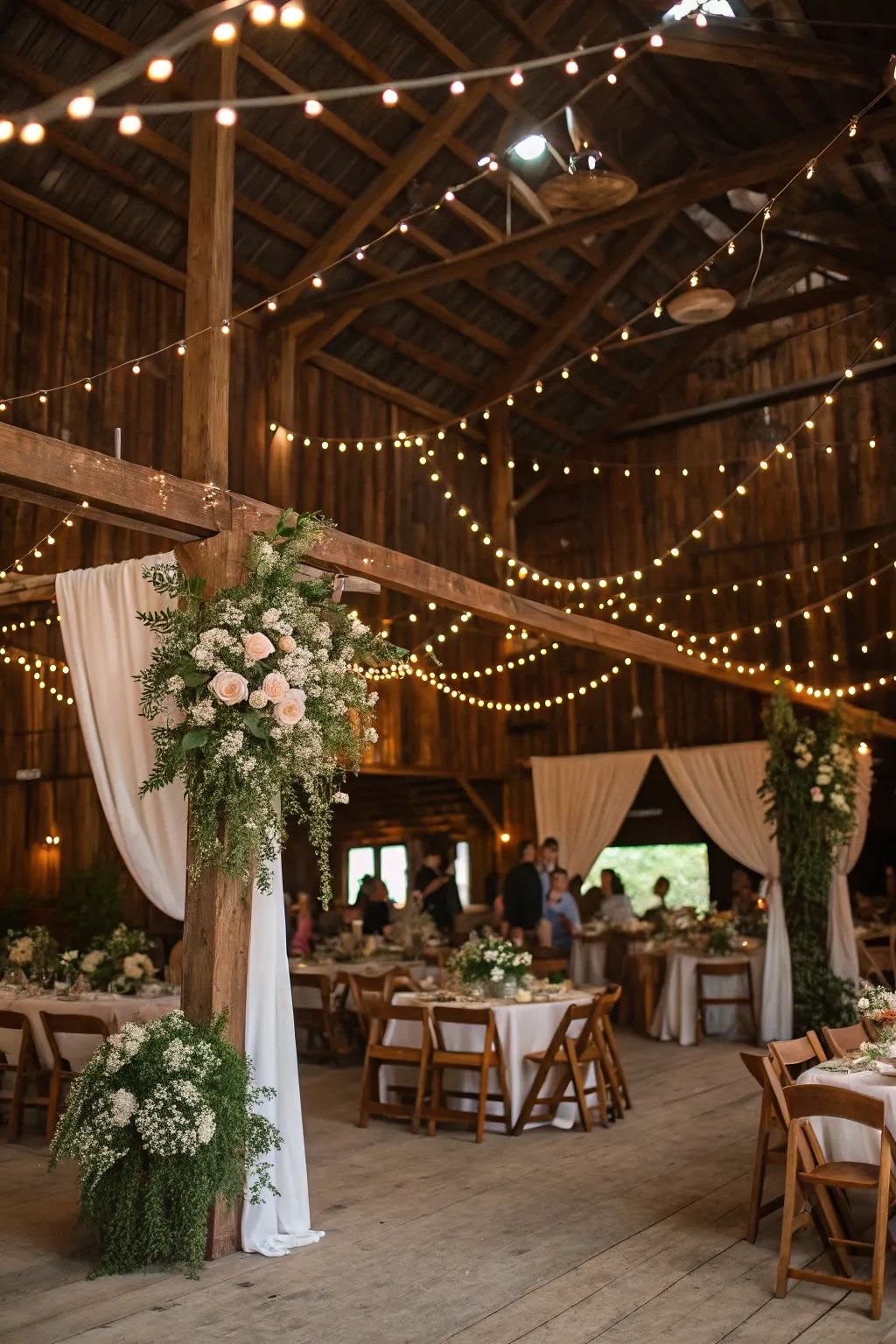 An indoor barn reception hall with round white tables, wooden cross-back chairs, and central timber posts wrapped in pink roses and green ivy.