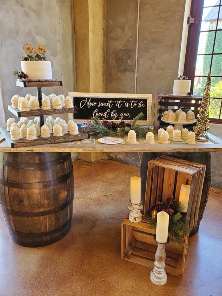 A rustic wedding cake display table supported by two wine barrels holding rows of mini tiered cakes and glowing floor candles.