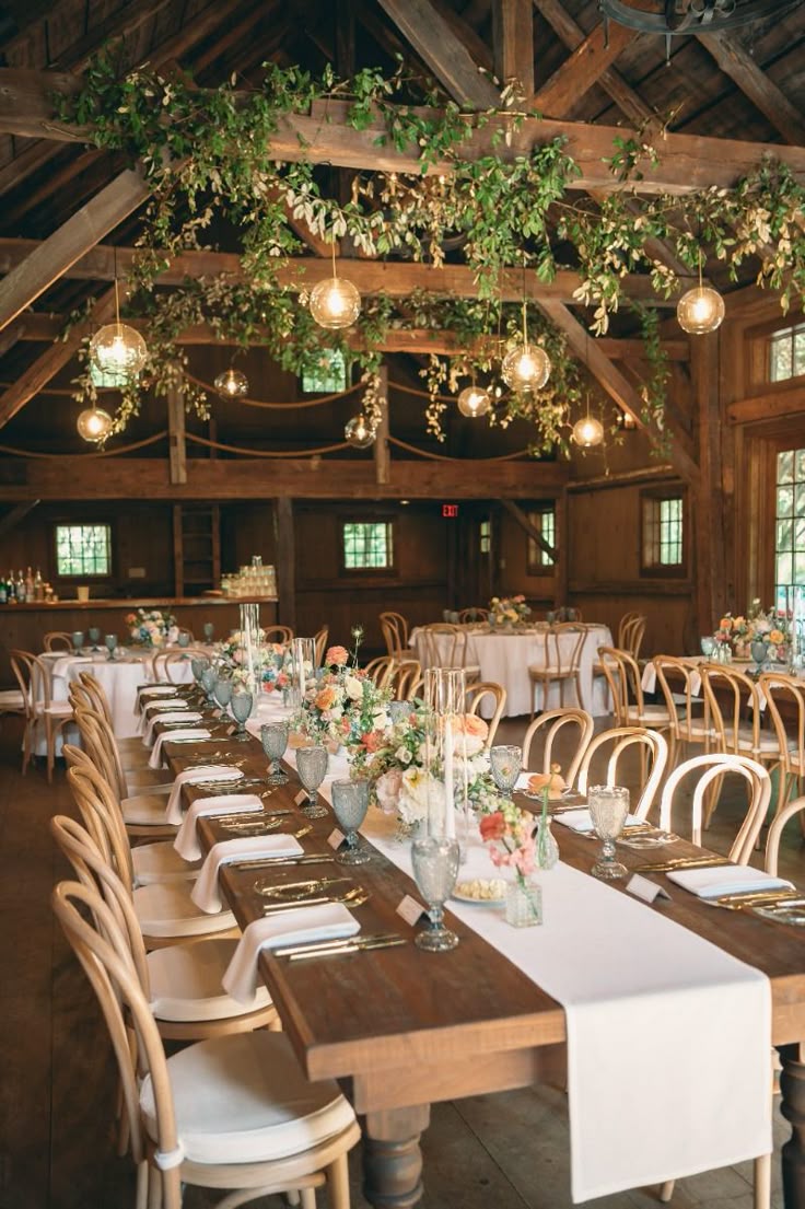 A long, indoor wooden banquet table with a white runner and floral centerpieces beneath a ceiling canopy of green foliage and round glass pendant lights.