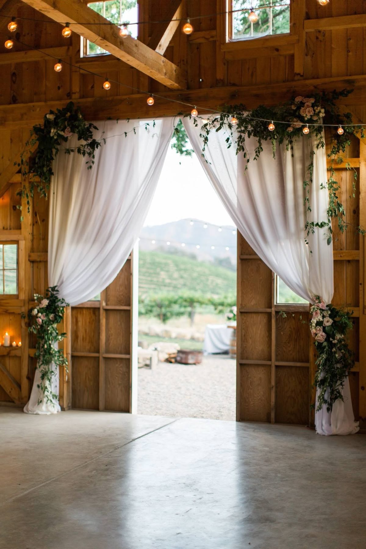 Open wooden barn doors framed by draped white curtains and rose clusters, looking out onto a scenic green vineyard path.