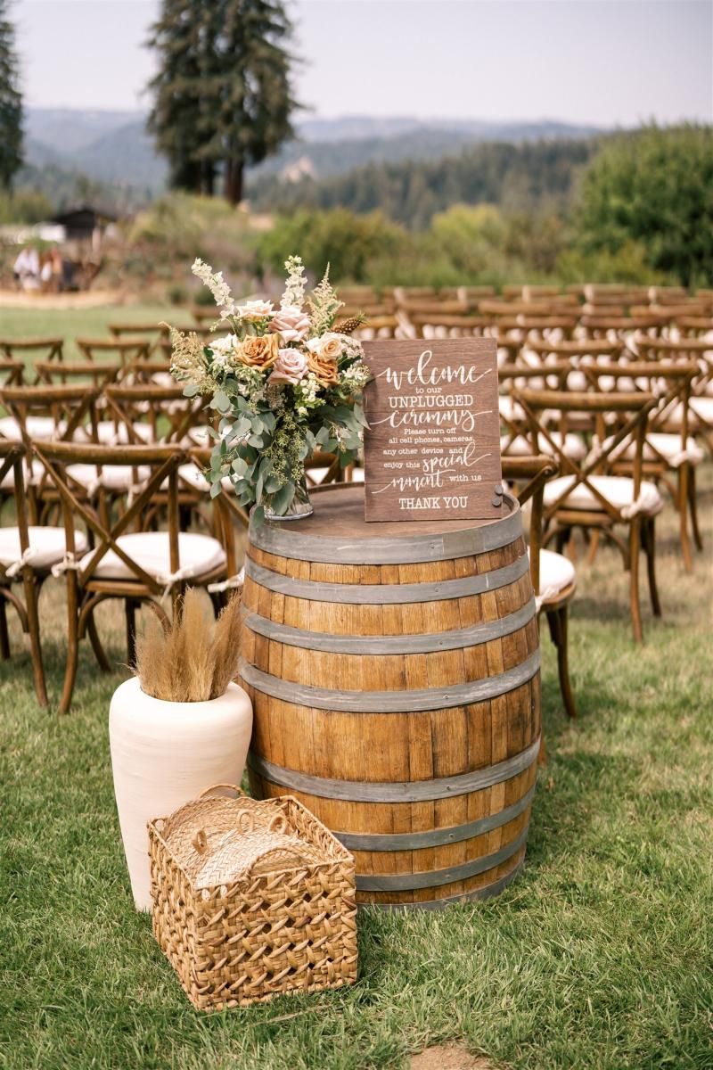 A rustic wooden unplugged ceremony sign sitting on a wine barrel with a basket and floral vase on a green lawn with wooden chairs.