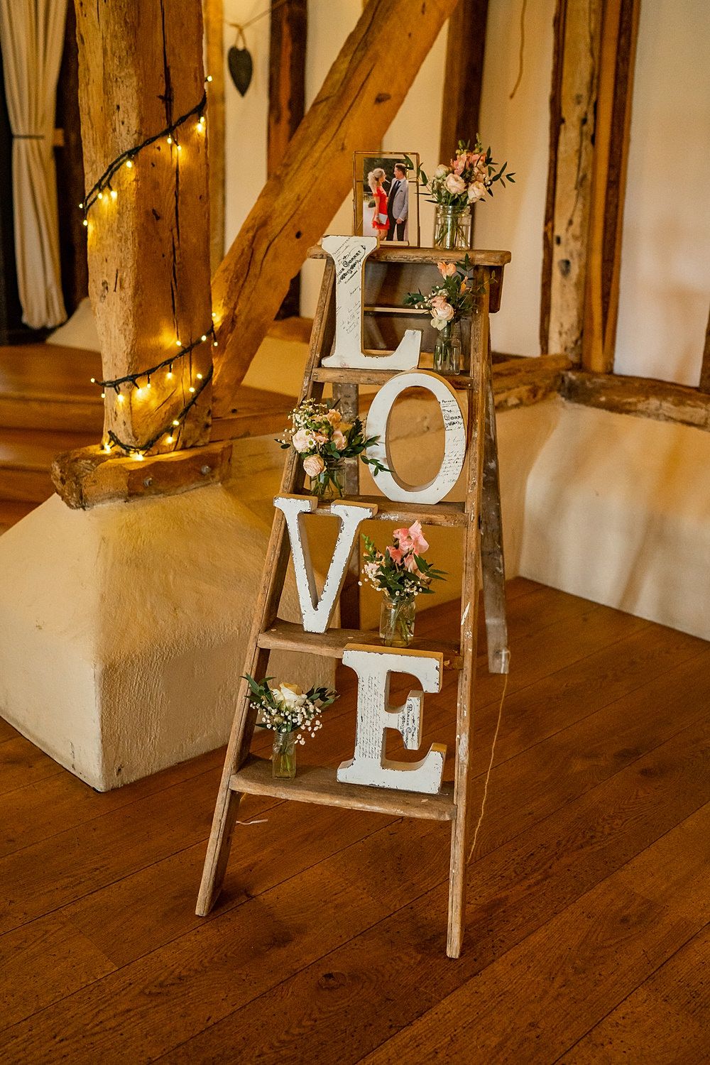 A rustic wooden stepladder holding large white letters spelling LOVE and small vases of pink flowers next to a lit timber column.