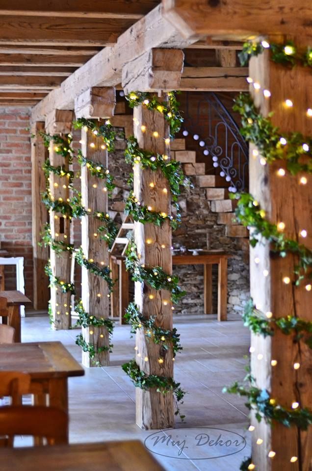 A row of indoor wooden support pillars wrapped vertically with green ivy garlands and warm twinkling fairy lights in a dining hall.
