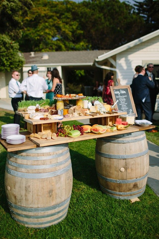 A multi-level grazing food station built on two wine barrels featuring cheeses, fruit, and bread on a grassy lawn with wedding guests behind.