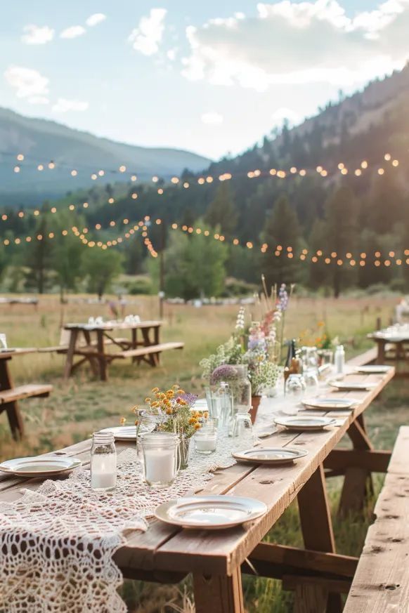 A long wooden outdoor picnic table set with lace runners, wildflower centerpieces, and candles under string lights with mountains in the background.