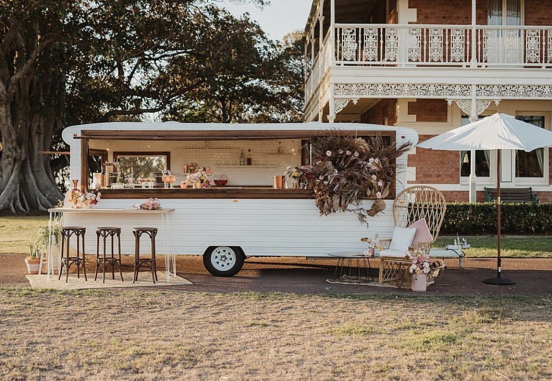 Elegant white caravan bar with timber counter, umbrella, peacock chair, and neutral dried florals.
