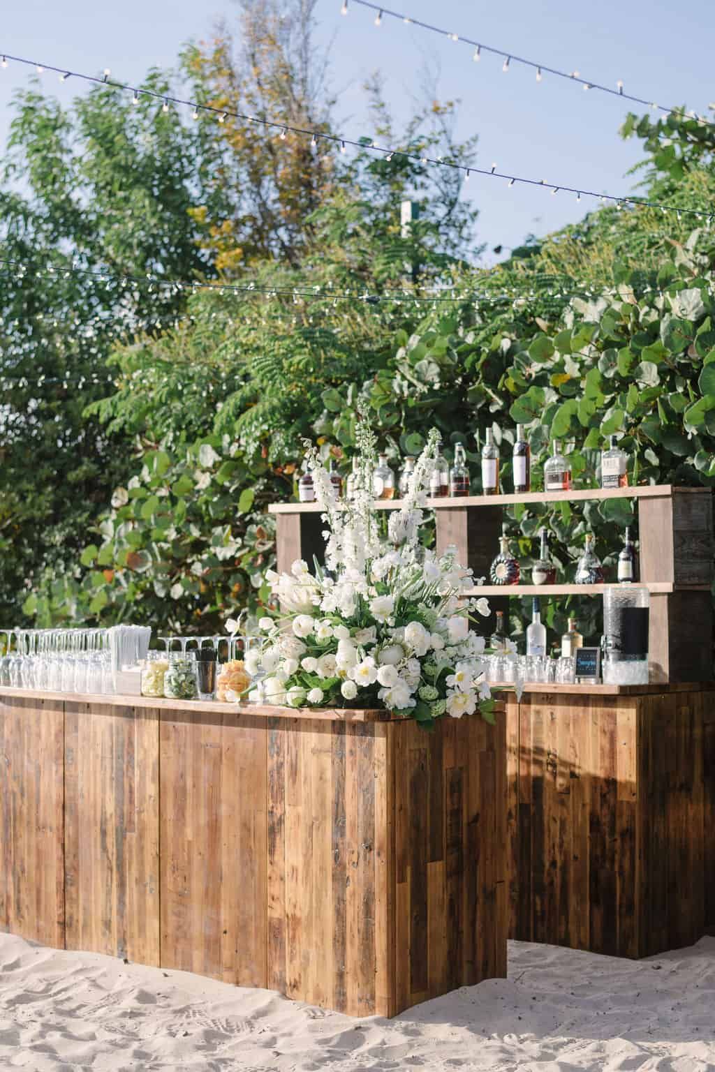 Rustic wooden outdoor bar setup on beach sand with tall white floral centerpiece and overhead string lights
