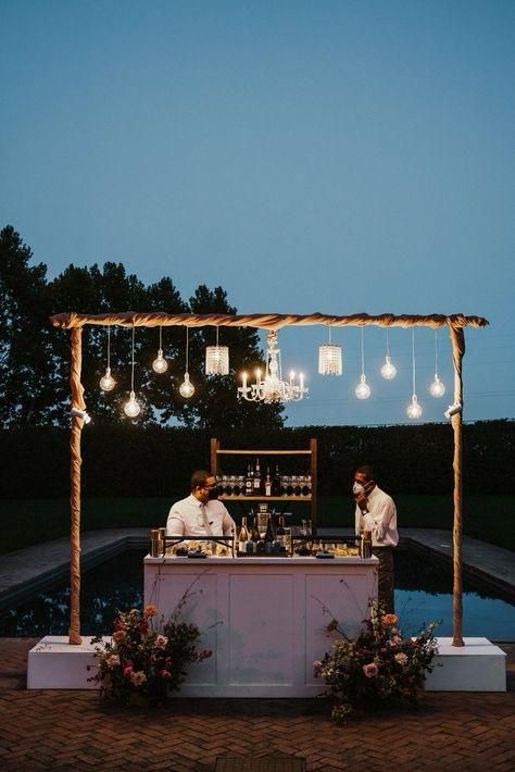 Modern white poolside bar with an overhead wooden frame suspending bistro bulbs and crystal chandeliers
