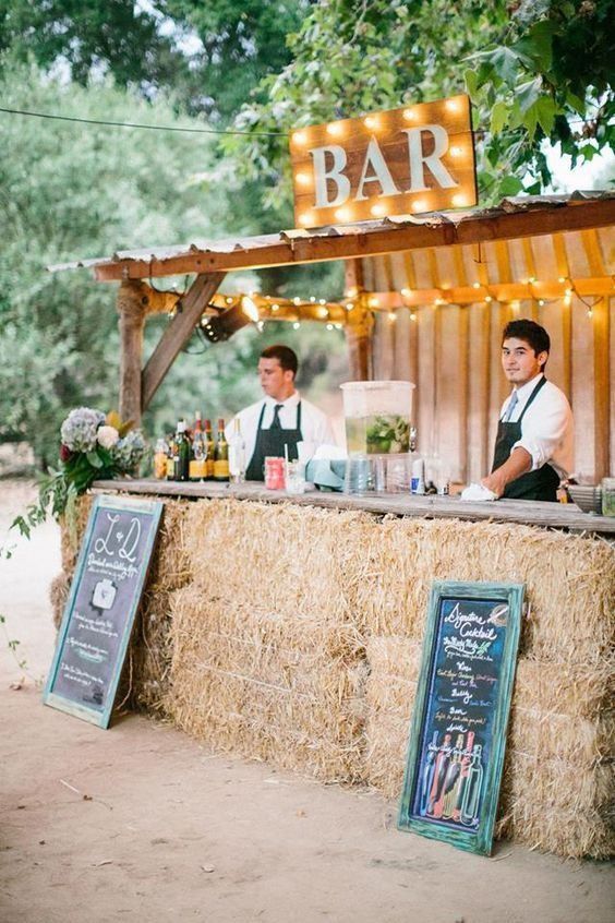 A wooden bar hut enclosed by stacked hay bales, featuring a marquee BAR sign and hanging chalkboard menus.
