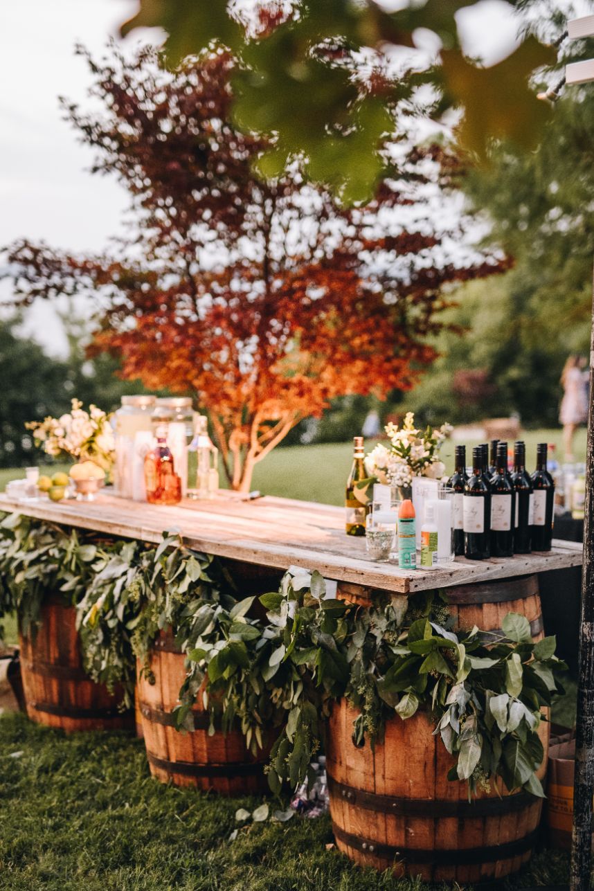 Wooden plank bar supported by wine barrels under a red maple tree, styled with leafy greenery garlands
