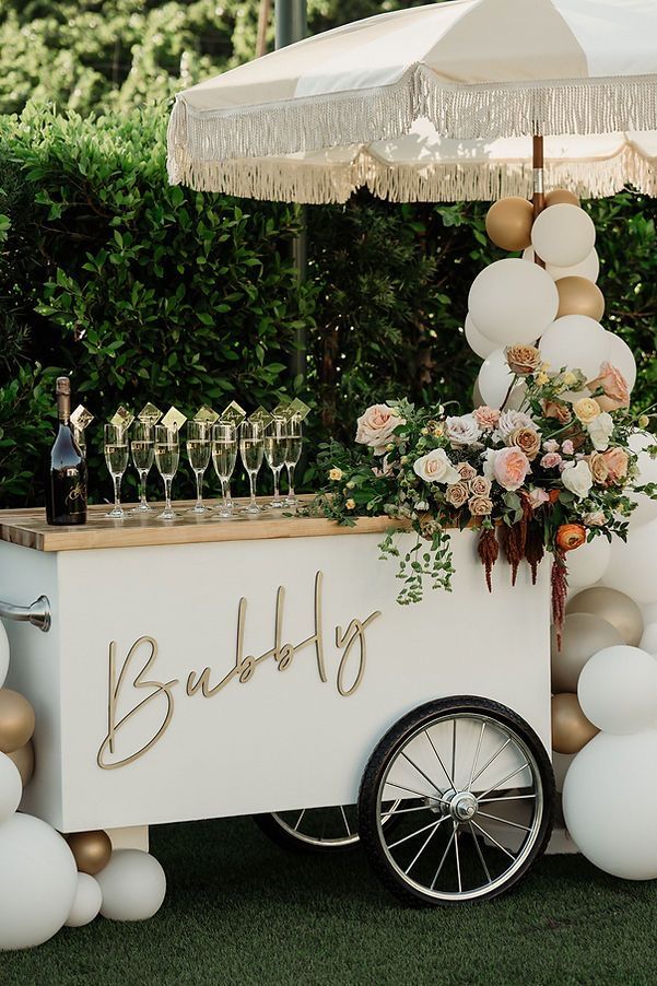 Retro white mobile champagne cart labeled Bubbly with a fringed umbrella, balloon garland, and fresh roses
