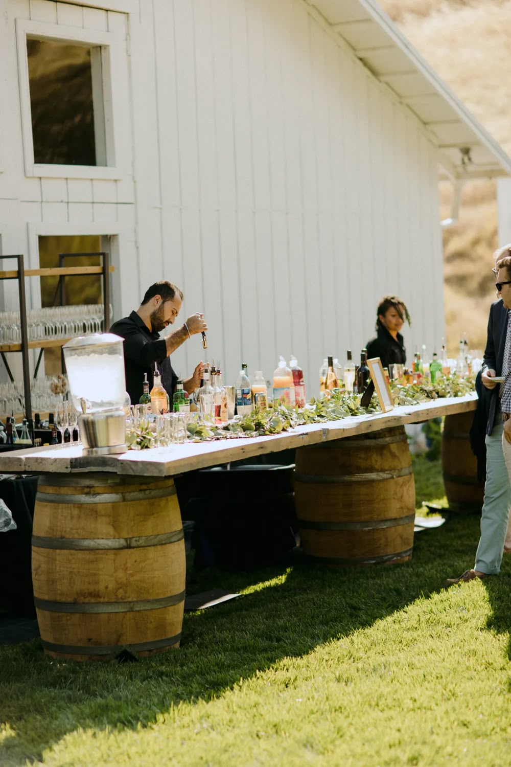 Long wooden bar on wine barrels placed on a lawn in front of a white barn

