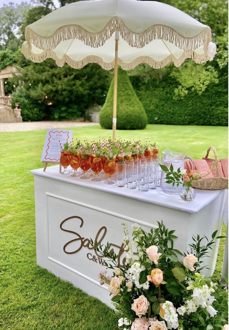 White rectangular bar top with a fringed market umbrella, pre-poured cocktails, and pink floral arrangement on the lawn
