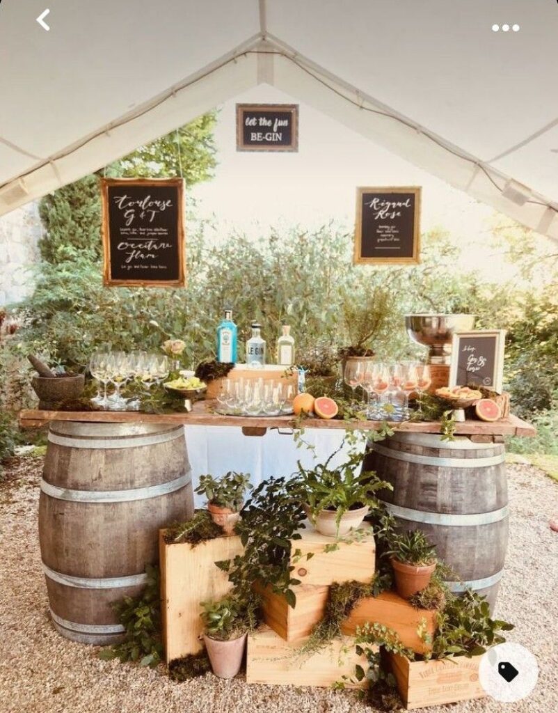 DIY gin bar station inside a tent with wine barrels, stacked wooden crates, potted plants, and chalkboard menus
