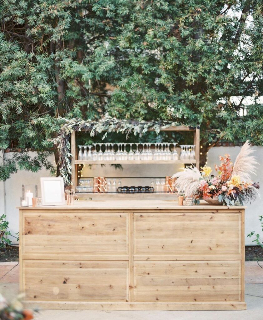 Light wood bar table styled with glass drink dispensers, tumblers, and potted fresh garden herbs against a hedge
