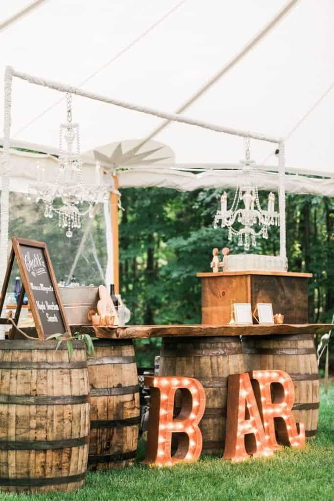 A wine barrel bar featuring a live-edge wood counter, marquee "BAR" letters, and hanging crystal chandeliers.
