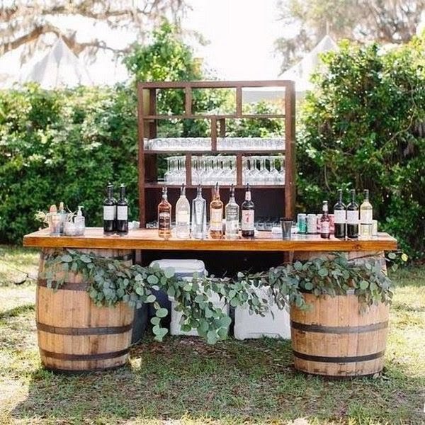 Outdoor bar made from two wine barrels with a eucalyptus garland and open wooden back shelving on grass.
