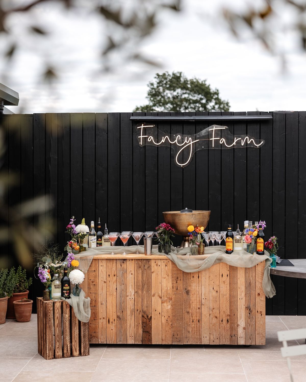 A natural wood plank bar staged against a black fence with sage green draping, colorful florals, and a light-up sign.
