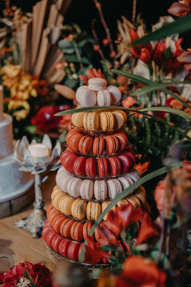 Tiered macaron tower in cream, blush, caramel, terracotta, and deep red shades surrounded by autumn flowers.