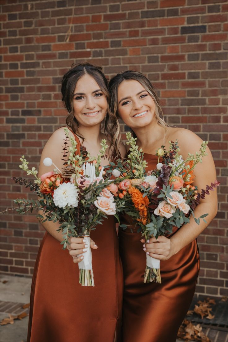 Two bridesmaids in rust satin dresses holding autumn bouquets of peach, orange, cream, and white flowers.