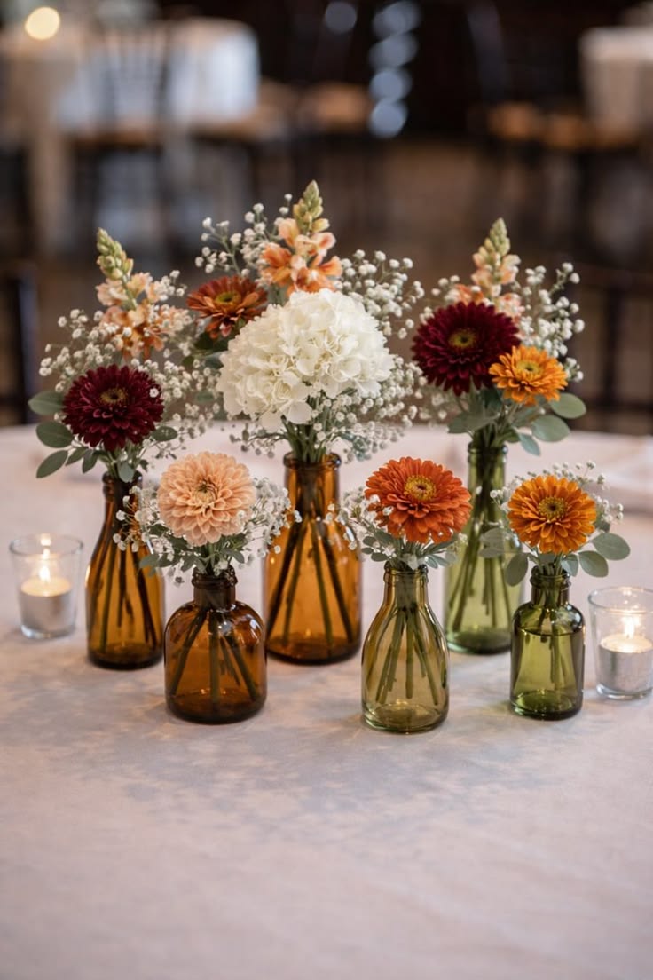Small amber and green glass bottles holding orange dahlias, peach flowers, white blooms, and greenery.